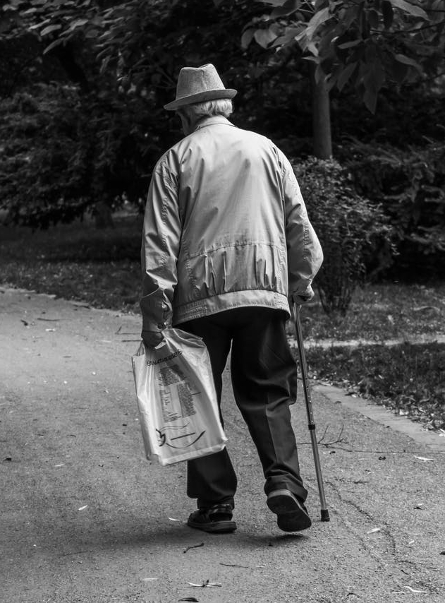 grayscale back view photo of elderly man with cane walking on dirt road
